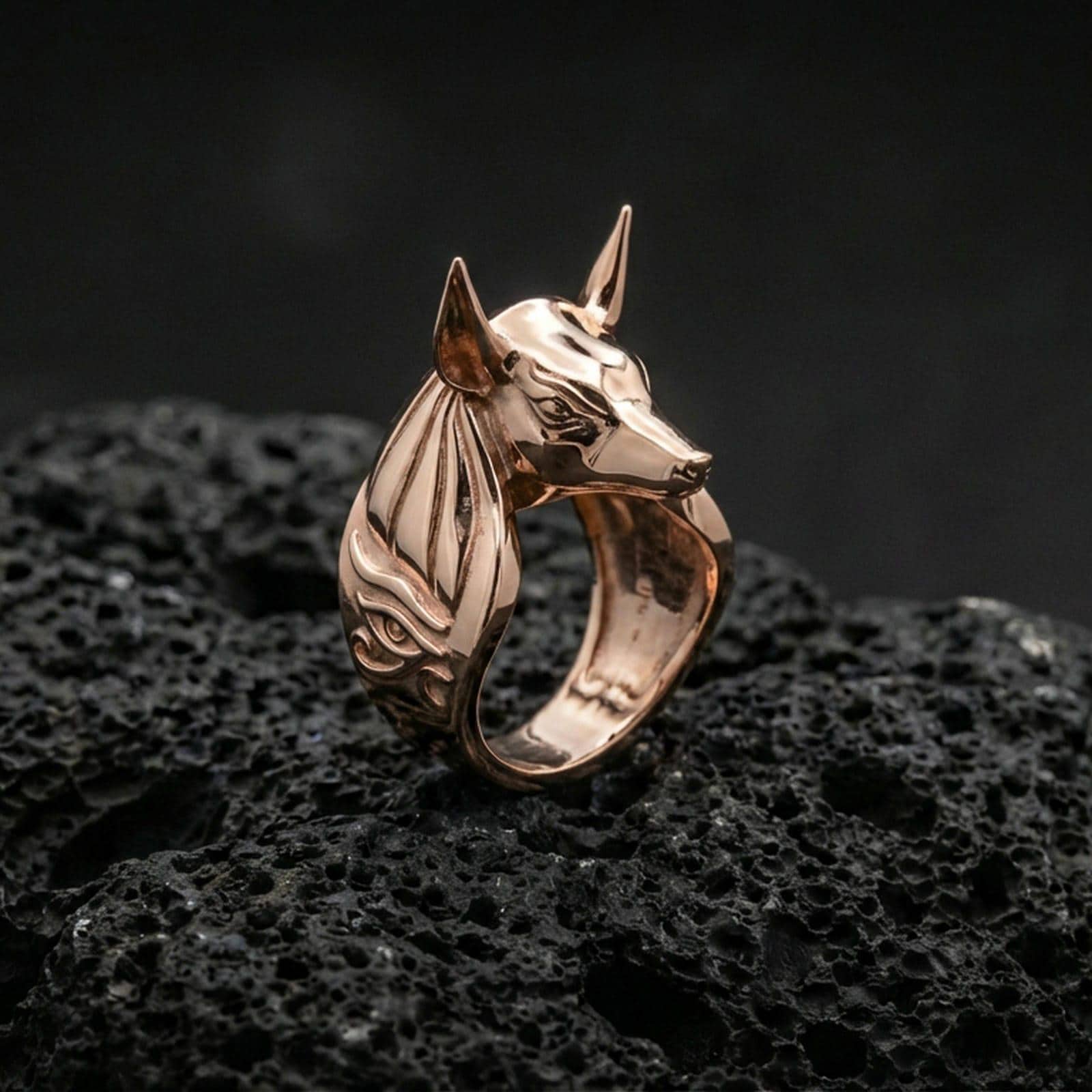 Anubis jackal head ring in a rose gold plated finish on volcanic rock, minimalist Egyptian mythology form with symbolic side motif, high-end close-up