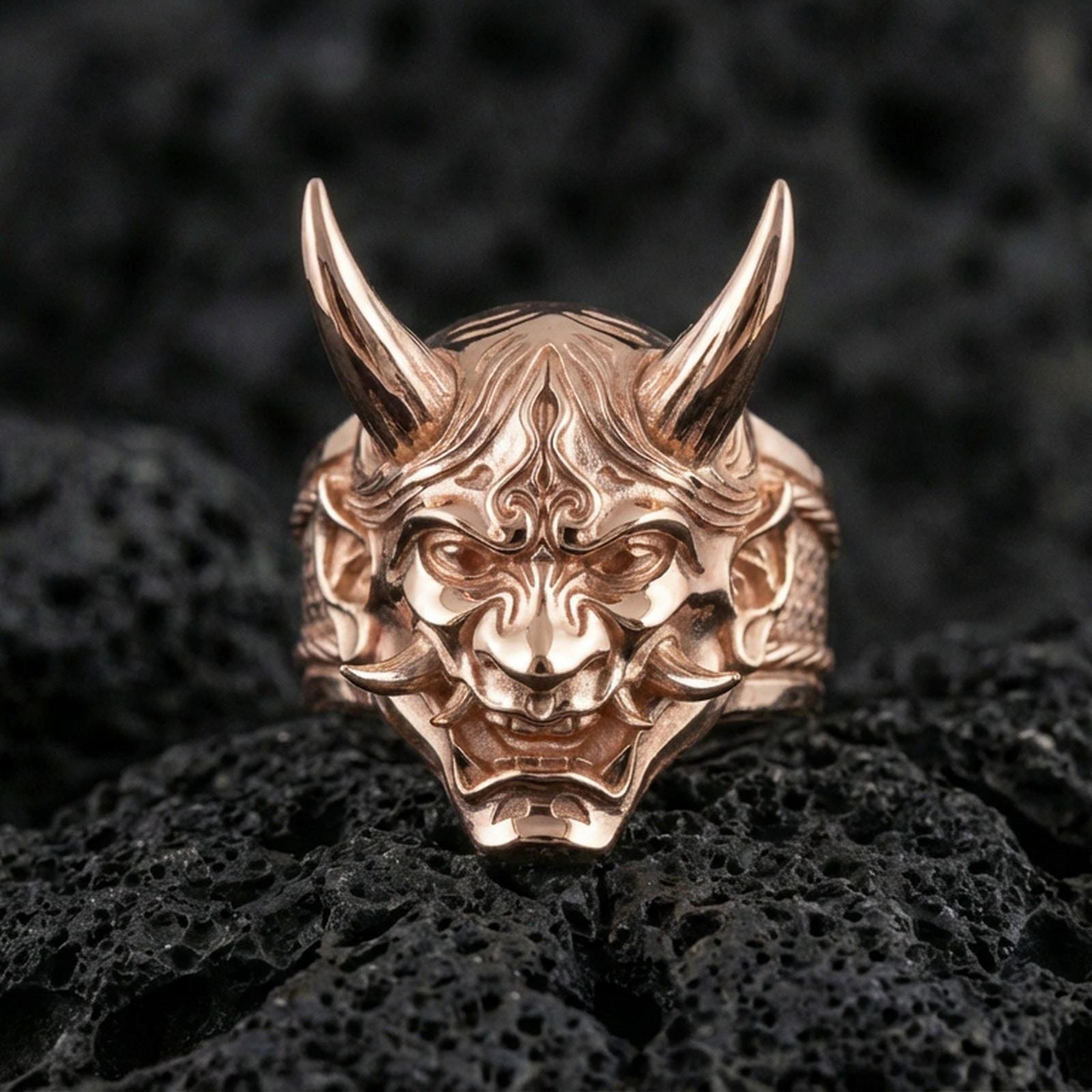 Sterling silver oni ring in a rose gold plated finish on volcanic rock, yokai demon mask design with sculpted horns and fangs, high-end close-up