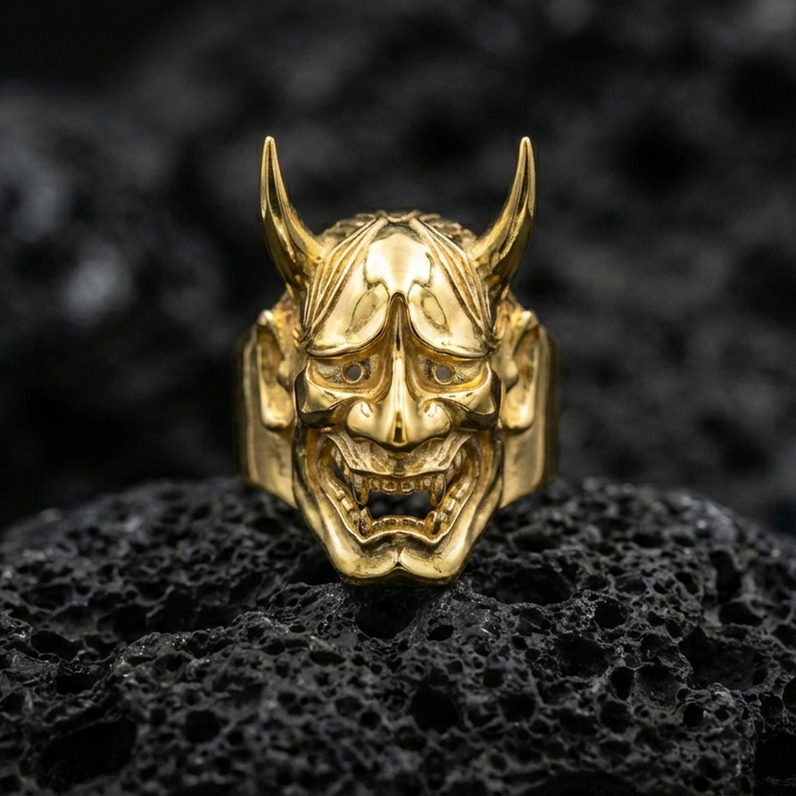 Japanese Hannya mask ring in a gold plated finish on volcanic rock, dramatic demon face carving with horns and teeth, luxury macro shot
