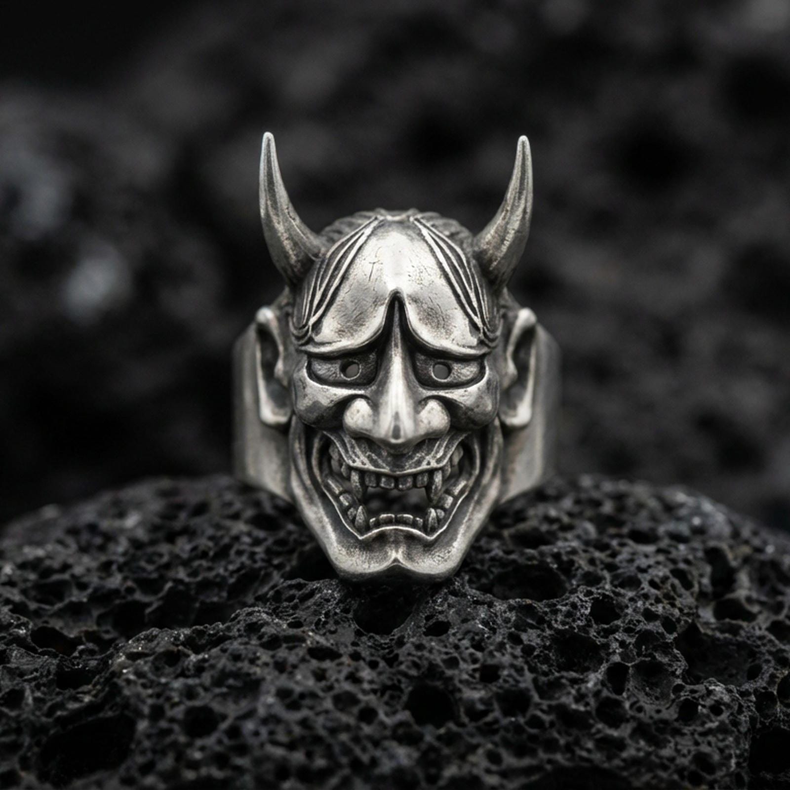 Sterling silver Hannya mask ring in an oxidized finish on volcanic rock, Japanese demon face with horns and grim expression, macro studio shot