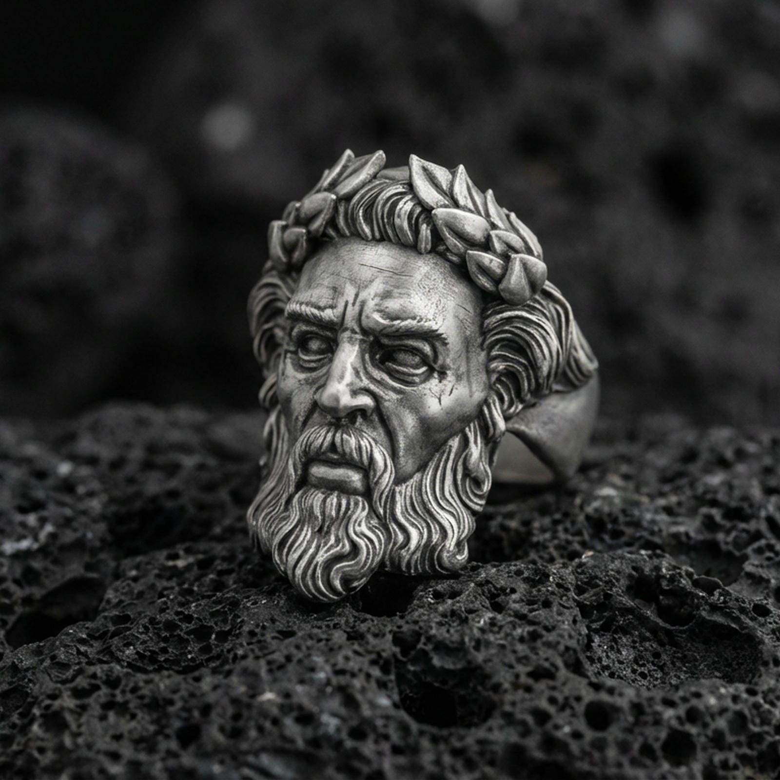 Detailed Zeus face ring in oxidized sterling silver with laurel wreath crown, macro product shot on textured volcanic rock background
