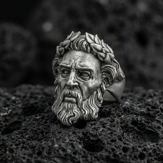 Detailed Zeus face ring in oxidized sterling silver with laurel wreath crown, macro product shot on textured volcanic rock background