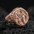 Load image into Gallery viewer, Rose gold plated griffon ring showcasing detailed winged beast engraving and refined sculptural texture, sharp close-up on volcanic rock
