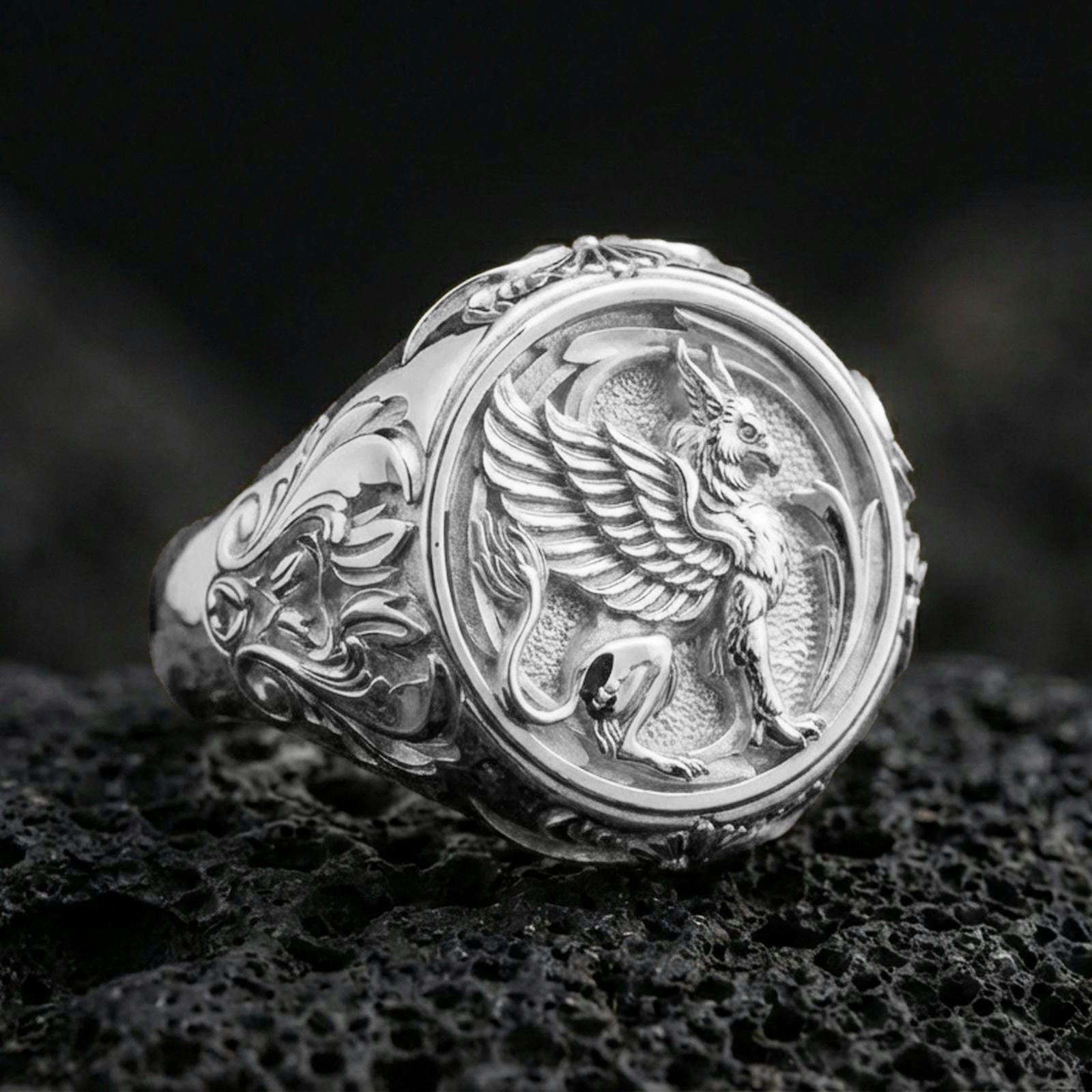 Polished silver griffon ring featuring sculpted griffin body and feathered wings, centered close-up jewelry photo on dark porous volcanic stone