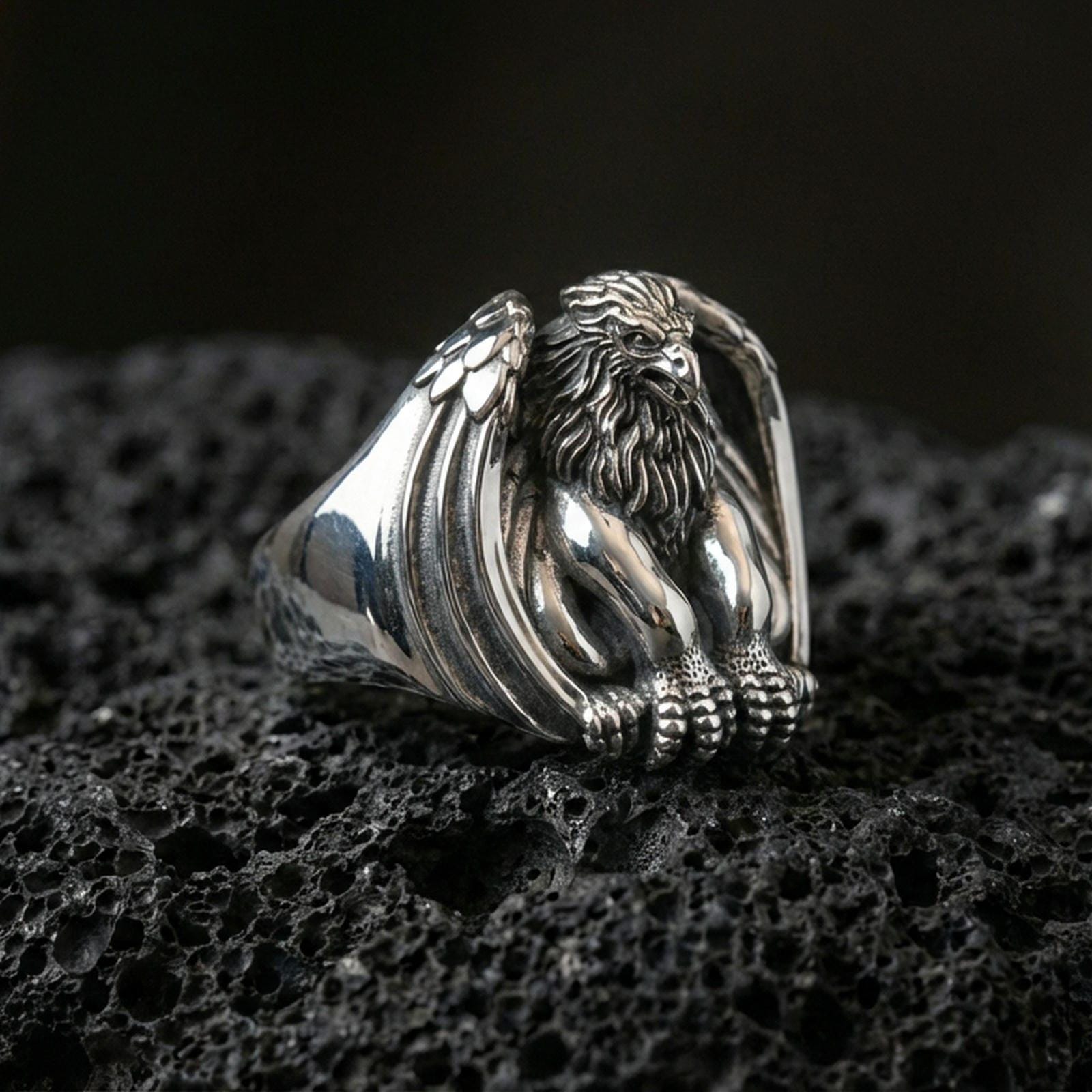 Polished silver griffin signet ring featuring guardian beast with folded wings and detailed claws, centered close-up on dark porous volcanic stone