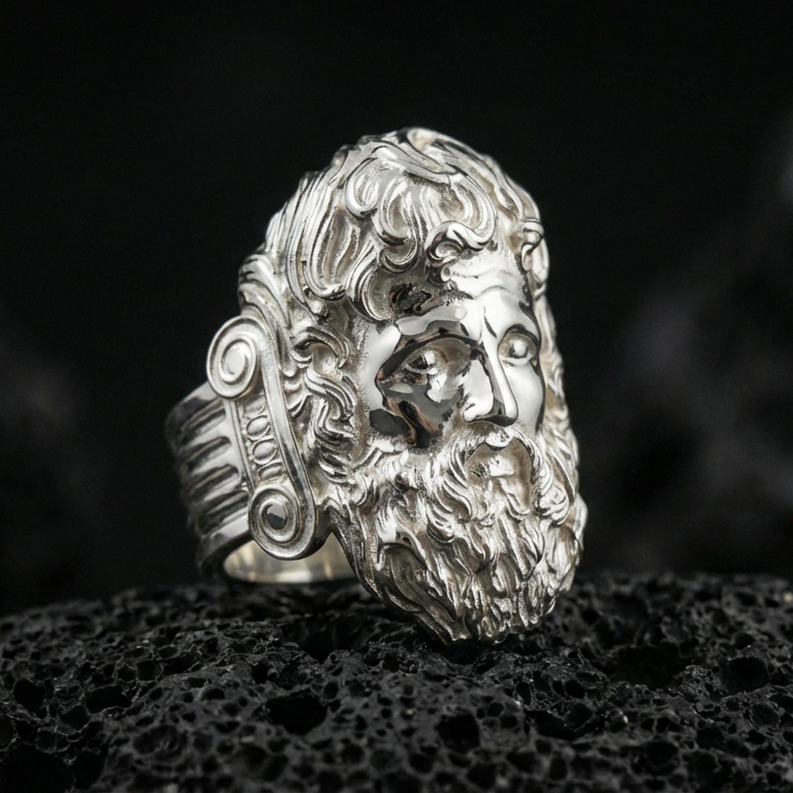 Polished silver Zeus ring featuring sculpted god portrait and refined facial expression, centered close-up jewelry photo on dark porous volcanic stone