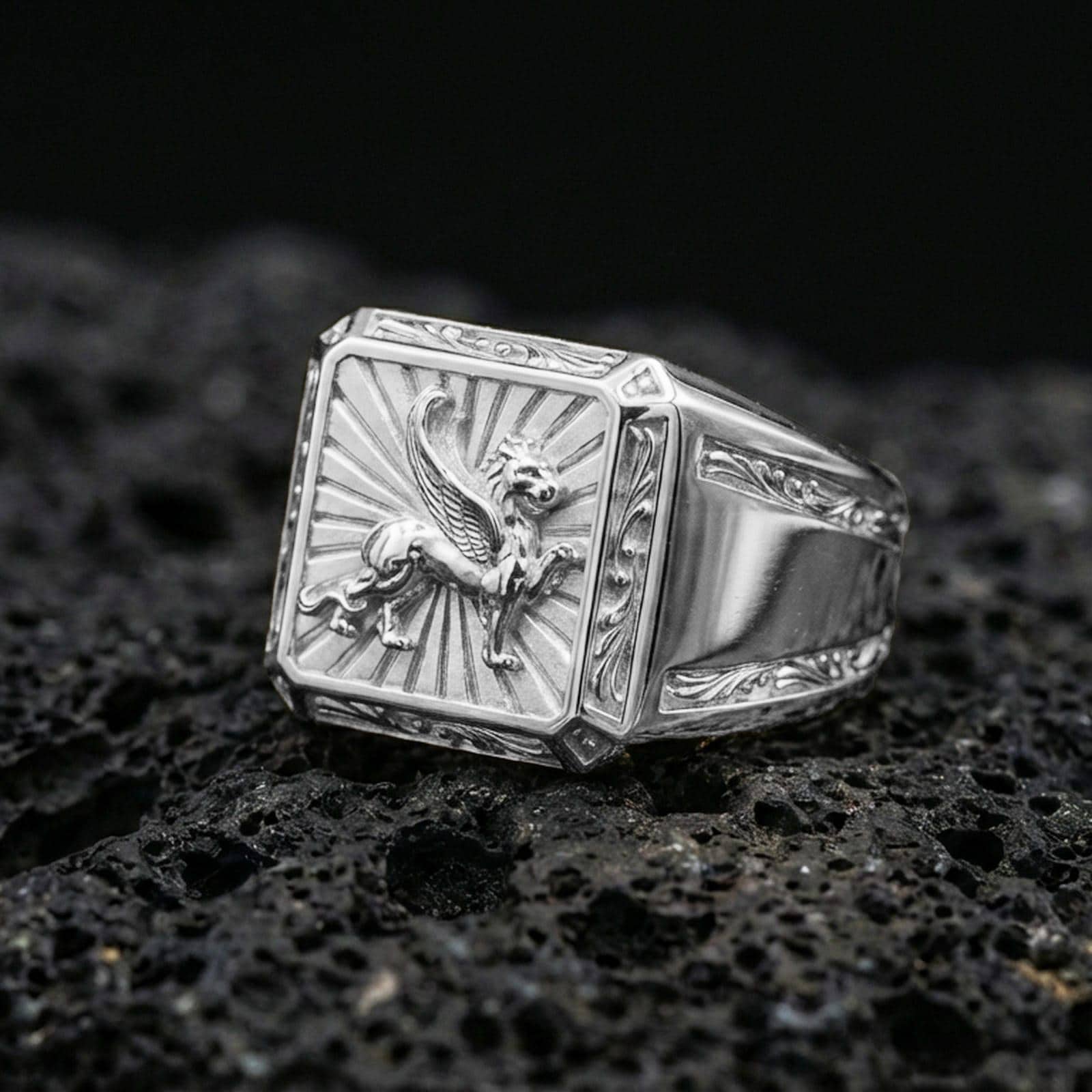 Polished silver gryphon ring featuring raised mythological beast relief and framed square signet design, centered close-up on dark porous volcanic stone