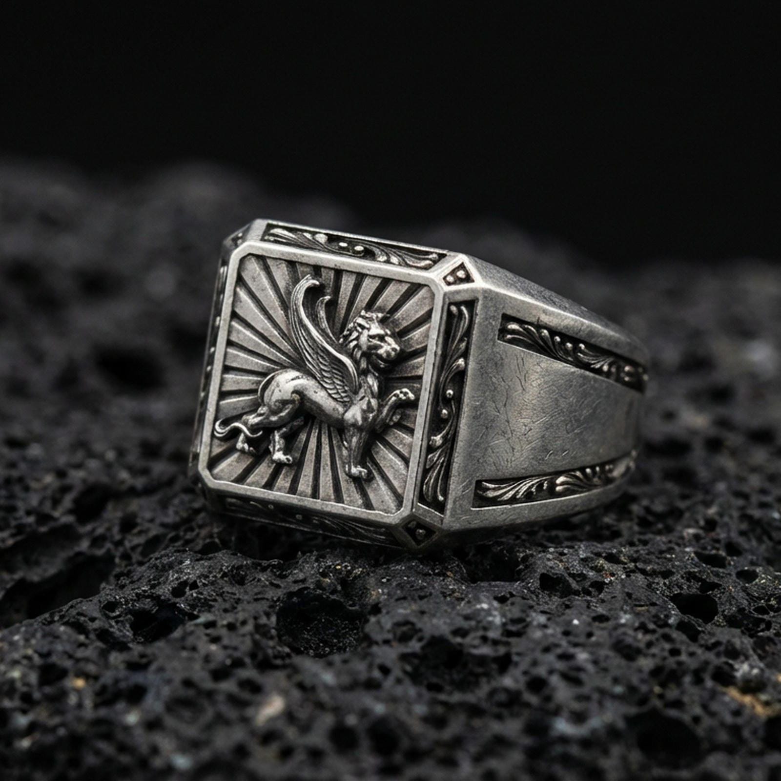 Detailed gryphon signet ring crafted in oxidized sterling silver, winged guardian creature emblem with sunburst background, macro product shot on textured volcanic rock