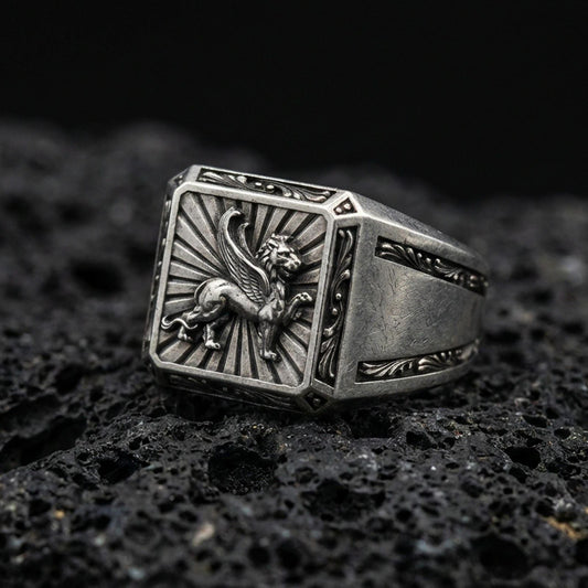 Detailed gryphon signet ring crafted in oxidized sterling silver, winged guardian creature emblem with sunburst background, macro product shot on textured volcanic rock