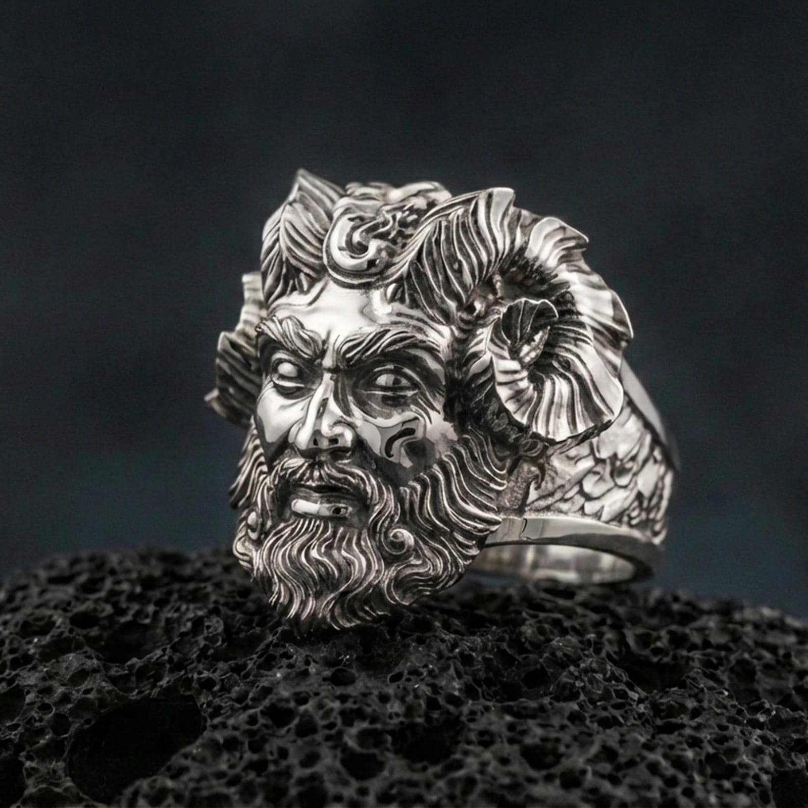 Polished silver Czernobog ring featuring sculpted horned god portrait and intricate hair texture, centered close-up on dark porous volcanic stone