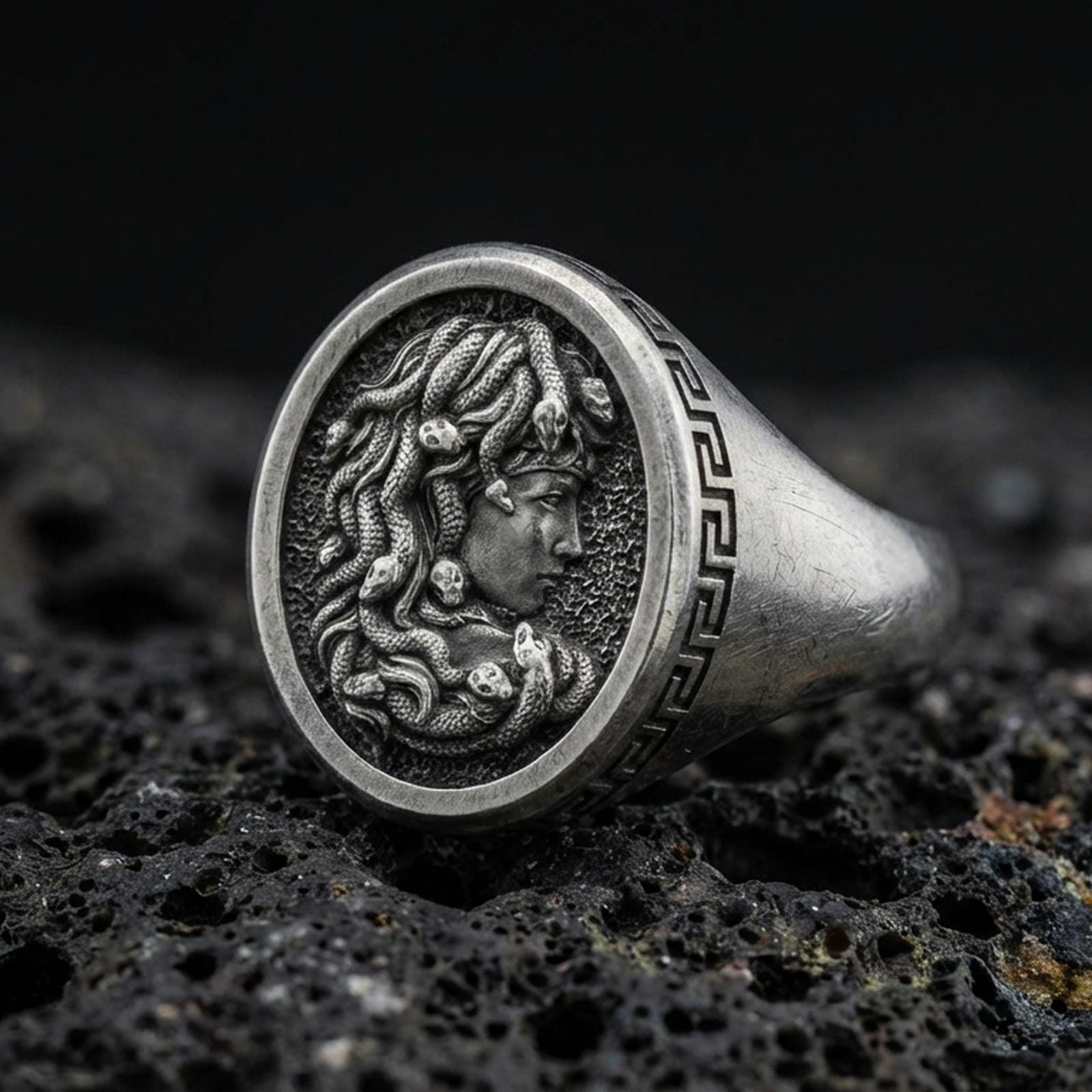 Detailed Medusa signet ring crafted in oxidized 925 sterling silver, snake hair portrait with Greek key border, macro product shot on textured volcanic rock background