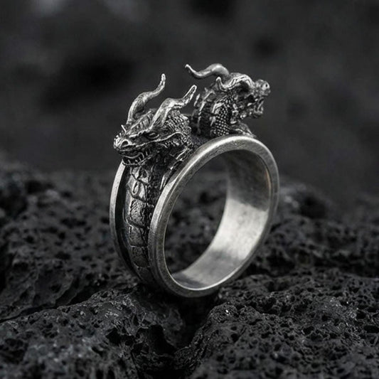 Detailed twin dragons ring crafted in oxidized sterling silver, two dragon heads meeting on an open band with scale texture, macro product shot on textured volcanic rock background