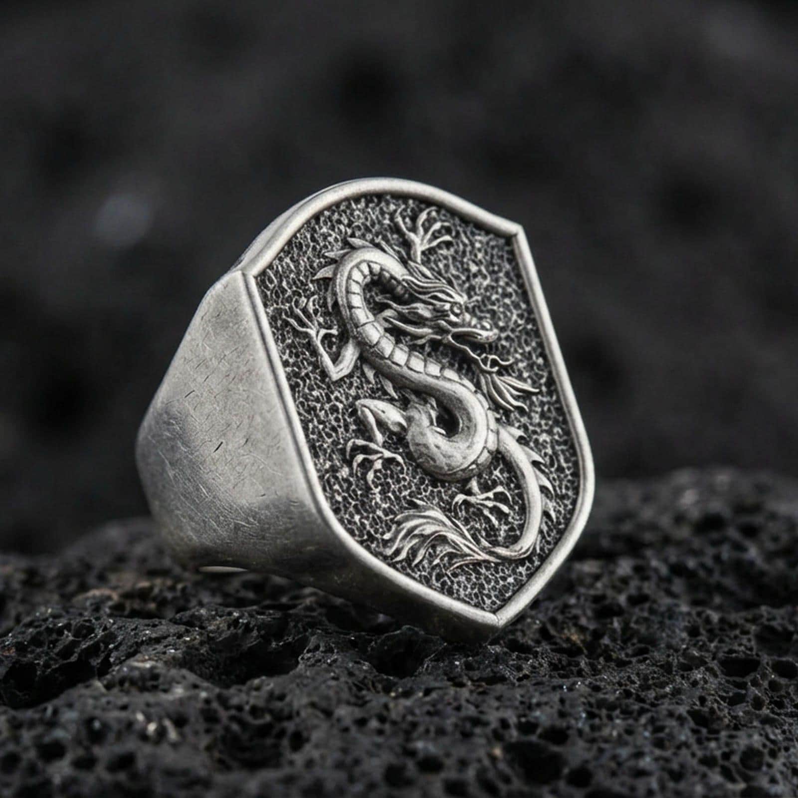 Bold Chinese dragon shield signet ring crafted in oxidized sterling silver, raised coiled dragon relief on textured signet face, macro product shot on volcanic rock background