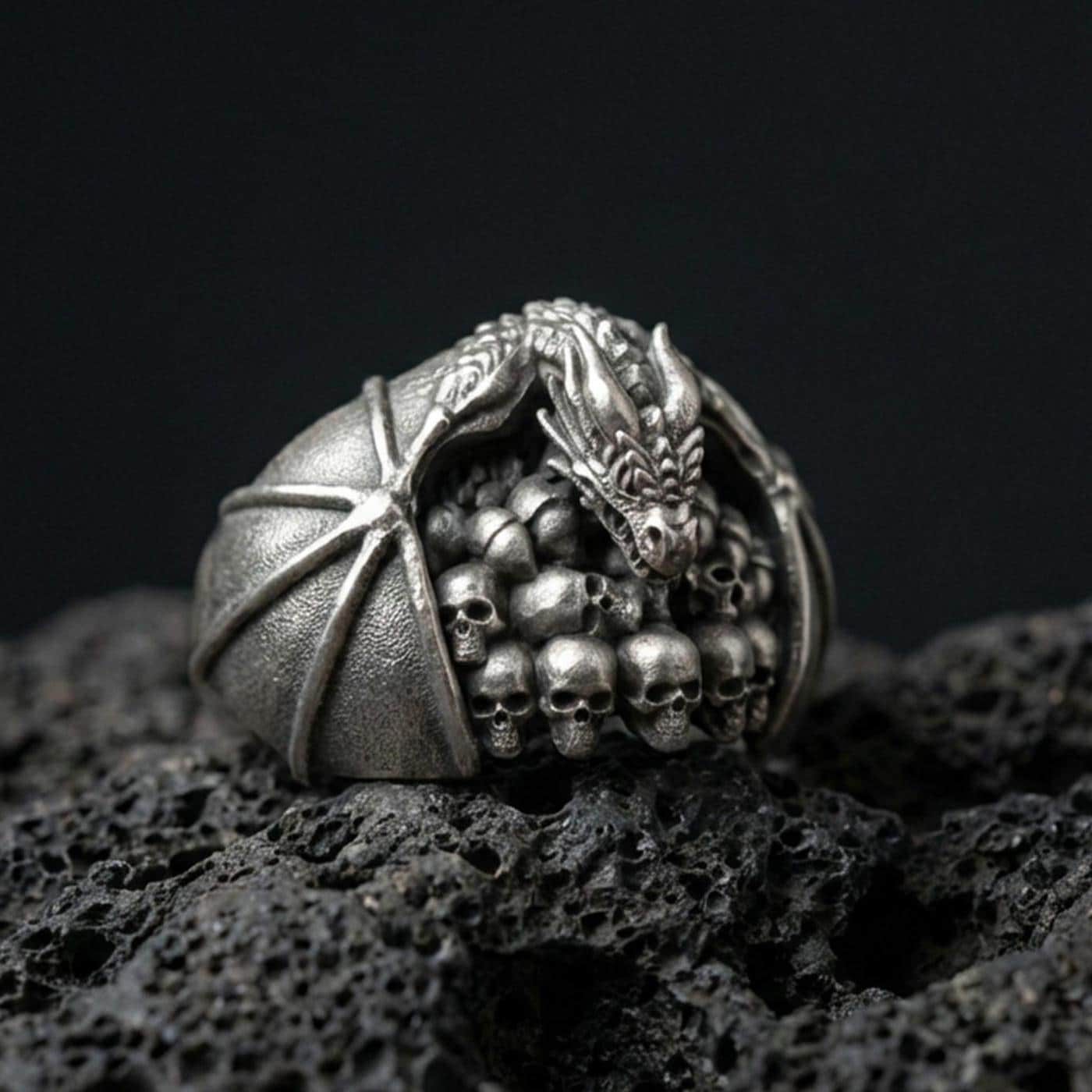 Oxidized sterling silver dragon skull ring on black volcanic stone, dragon perched over skull pile, gothic fantasy signet