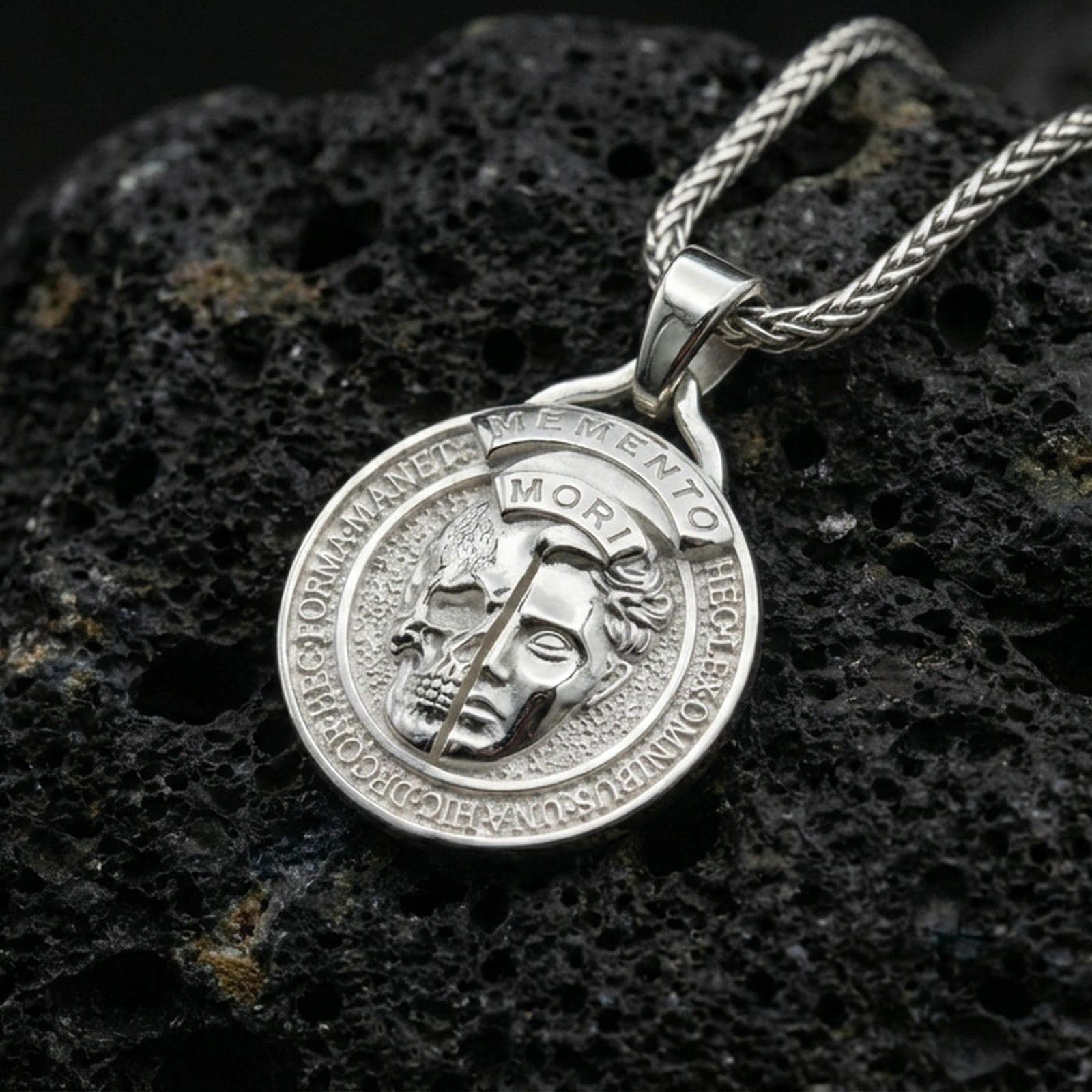 Polished silver memento mori medallion necklace on dark volcanic rock, Latin border engraving detail, premium close-up