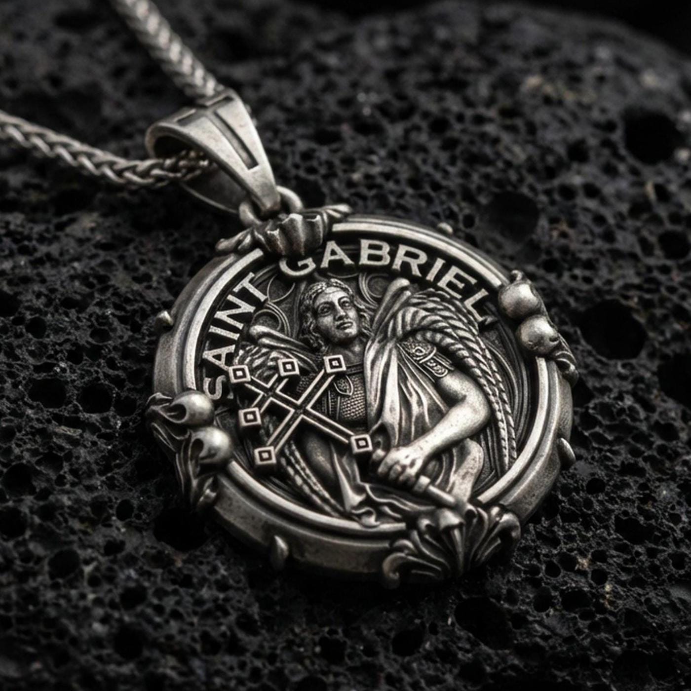 Oxidized sterling silver Saint Gabriel pendant on textured volcanic rock, archangel relief with cross motif, macro studio lighting