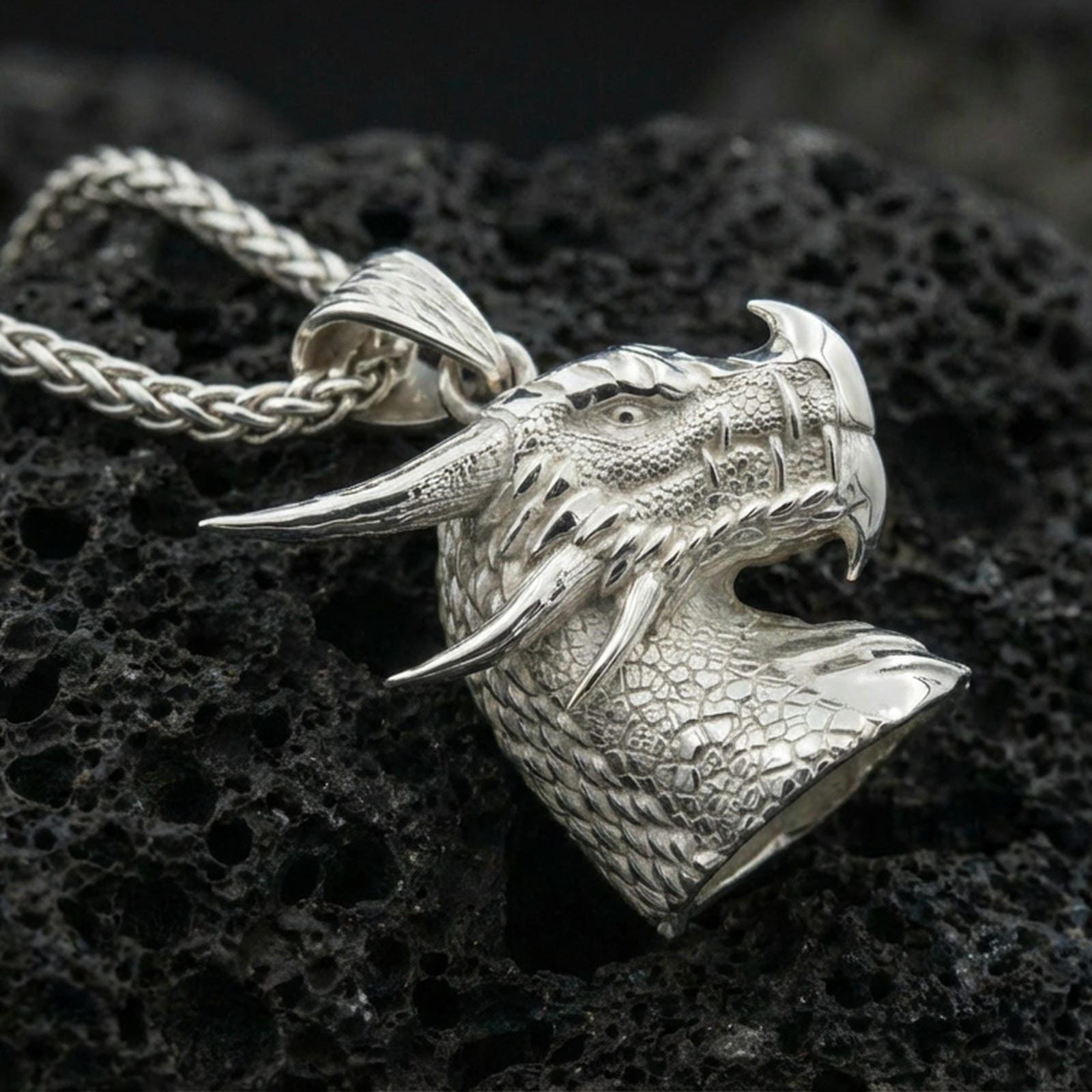 Polished silver dragon head pendant on dark volcanic stone, sharp fang and scale relief, premium close-up composition