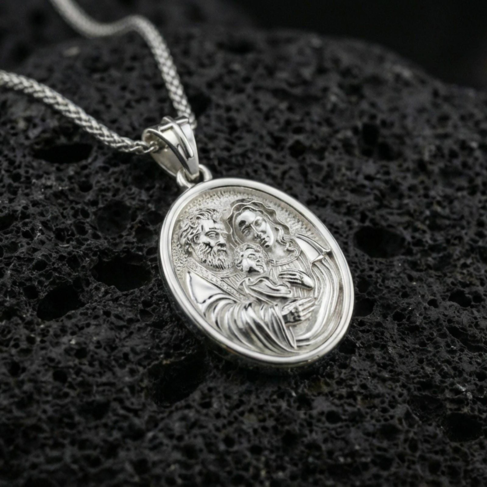 Polished silver Holy Family medallion necklace on dark volcanic stone, crisp engraved faces and smooth oval bezel, premium close-up composition