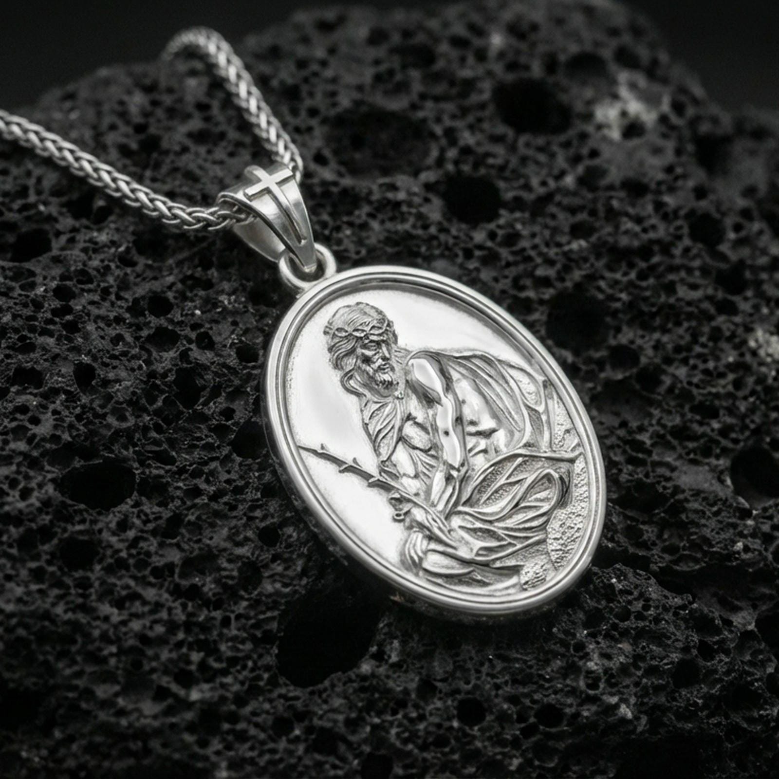 Polished silver Jesus Christ medallion necklace on dark volcanic stone, crisp engraved figure and smooth oval bezel, premium close-up composition
