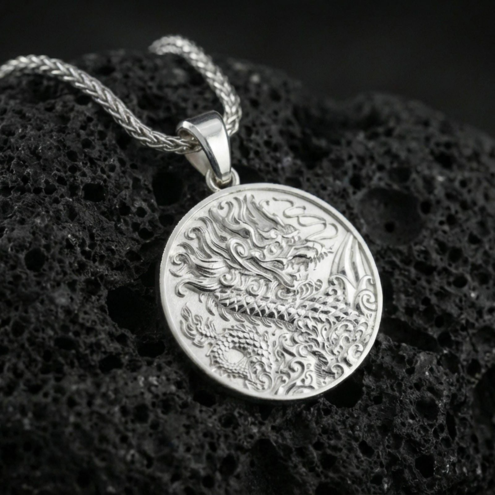 Polished silver Chinese dragon coin pendant necklace on dark volcanic stone, crisp scale relief and swirling cloud details, premium close-up composition