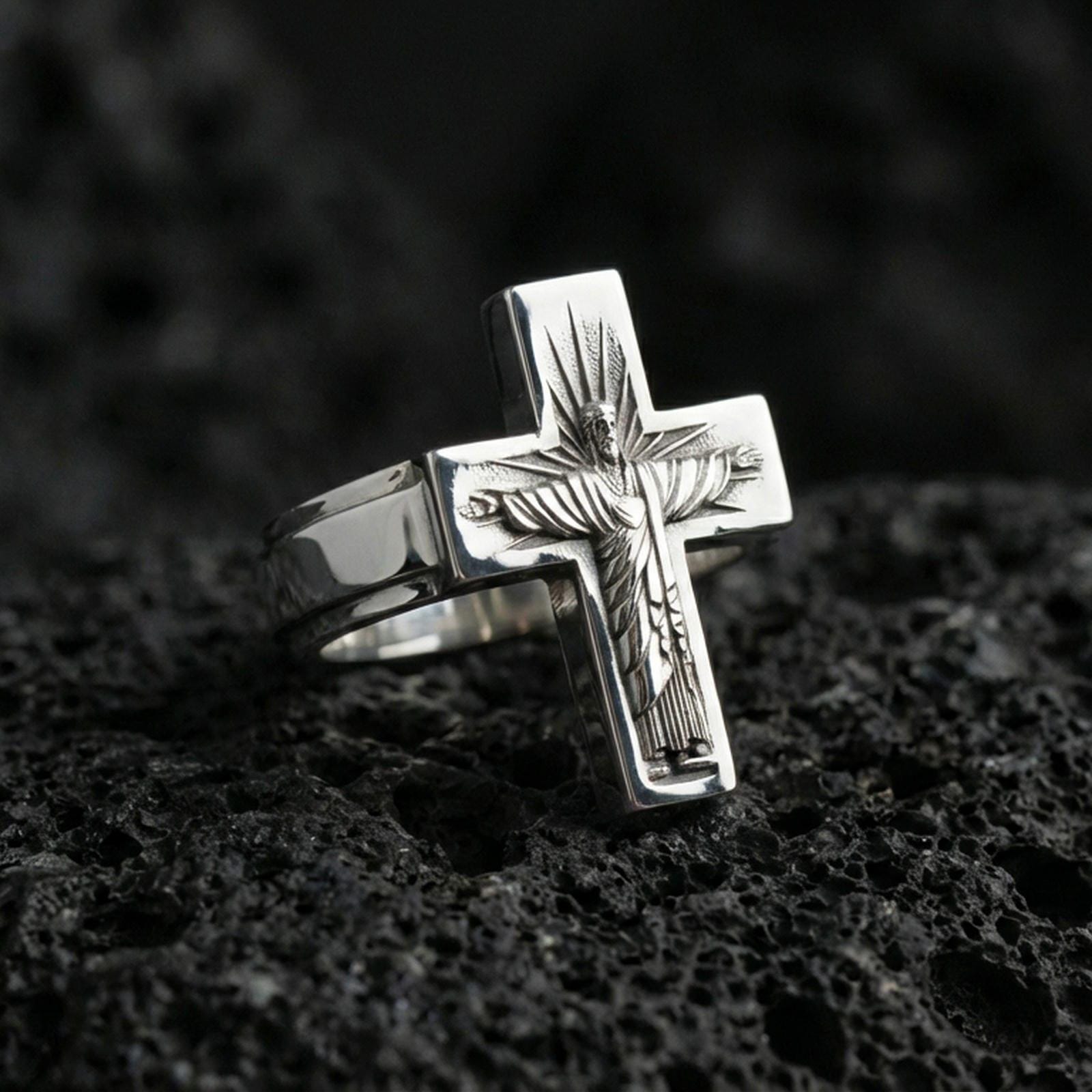 Polished silver Jesus crucifix ring on porous volcanic stone, detailed Christ figure engraving, bold cross silhouette ring
