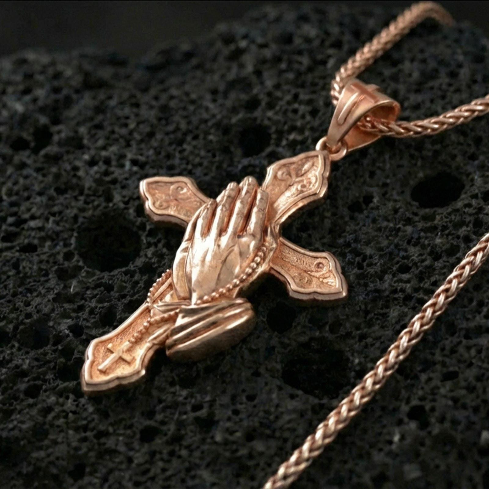 Rose gold plated praying hands cross pendant on textured volcanic rock, luxury close-up capturing the sculpted hands, rosary beads, and warm metallic glow