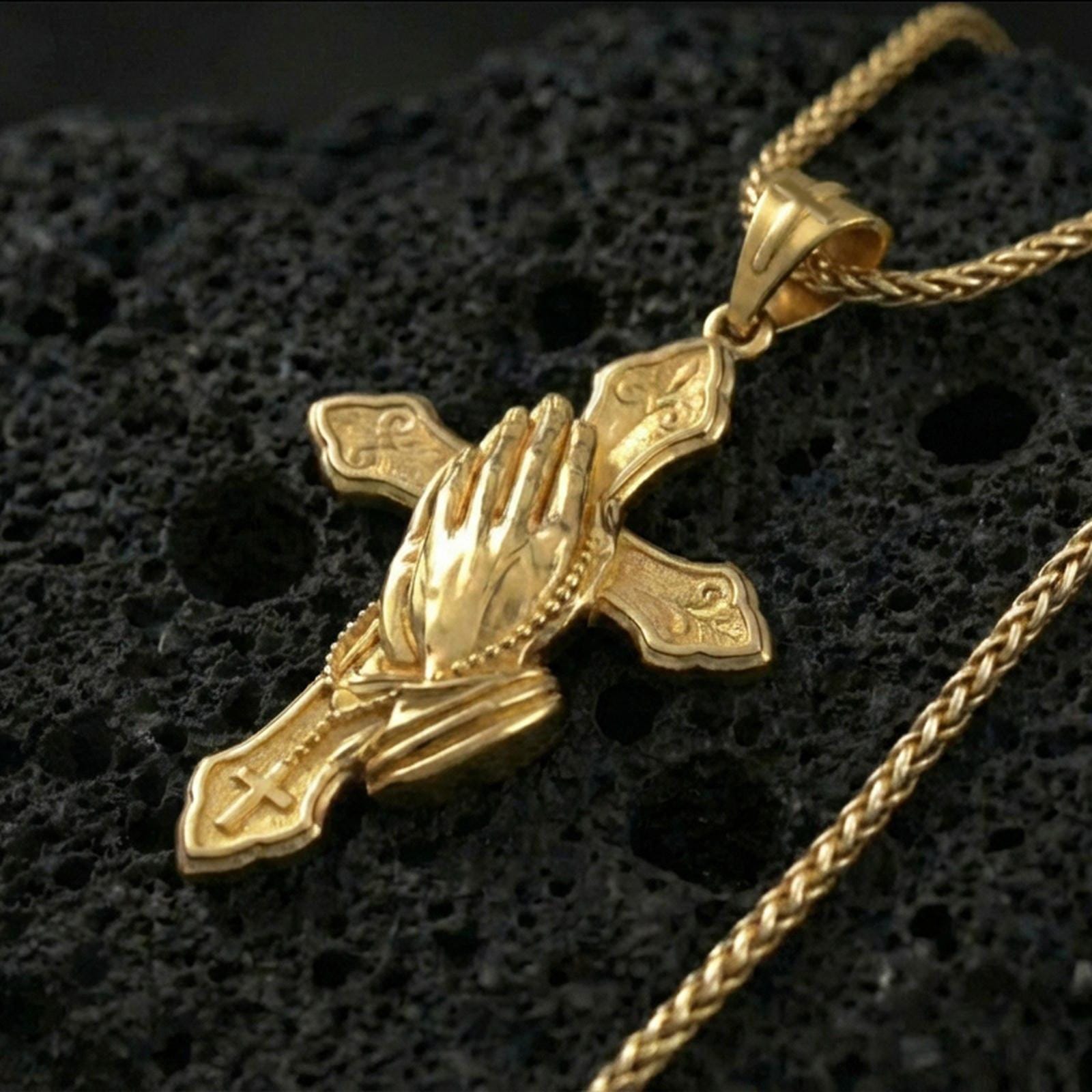 Gold plated praying hands cross pendant necklace on volcanic rock, premium macro shot emphasizing the rosary chain relief, engraved cross shapes, and bright shine