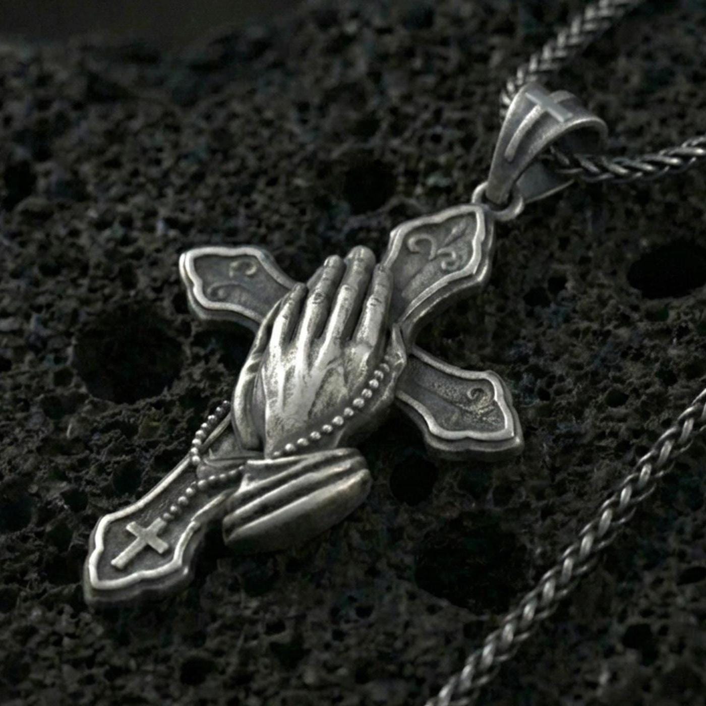 Oxidized sterling silver praying hands cross pendant necklace on black volcanic rock, macro close-up showing the rosary beads, cuff detail, and antique depth