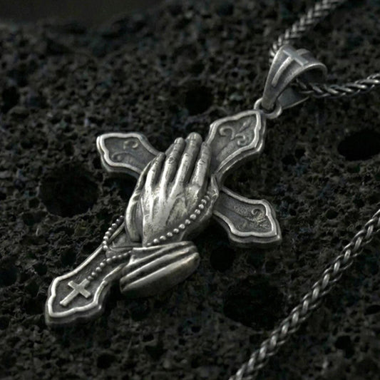 Oxidized sterling silver praying hands cross pendant necklace on black volcanic rock, macro close-up showing the rosary beads, cuff detail, and antique depth