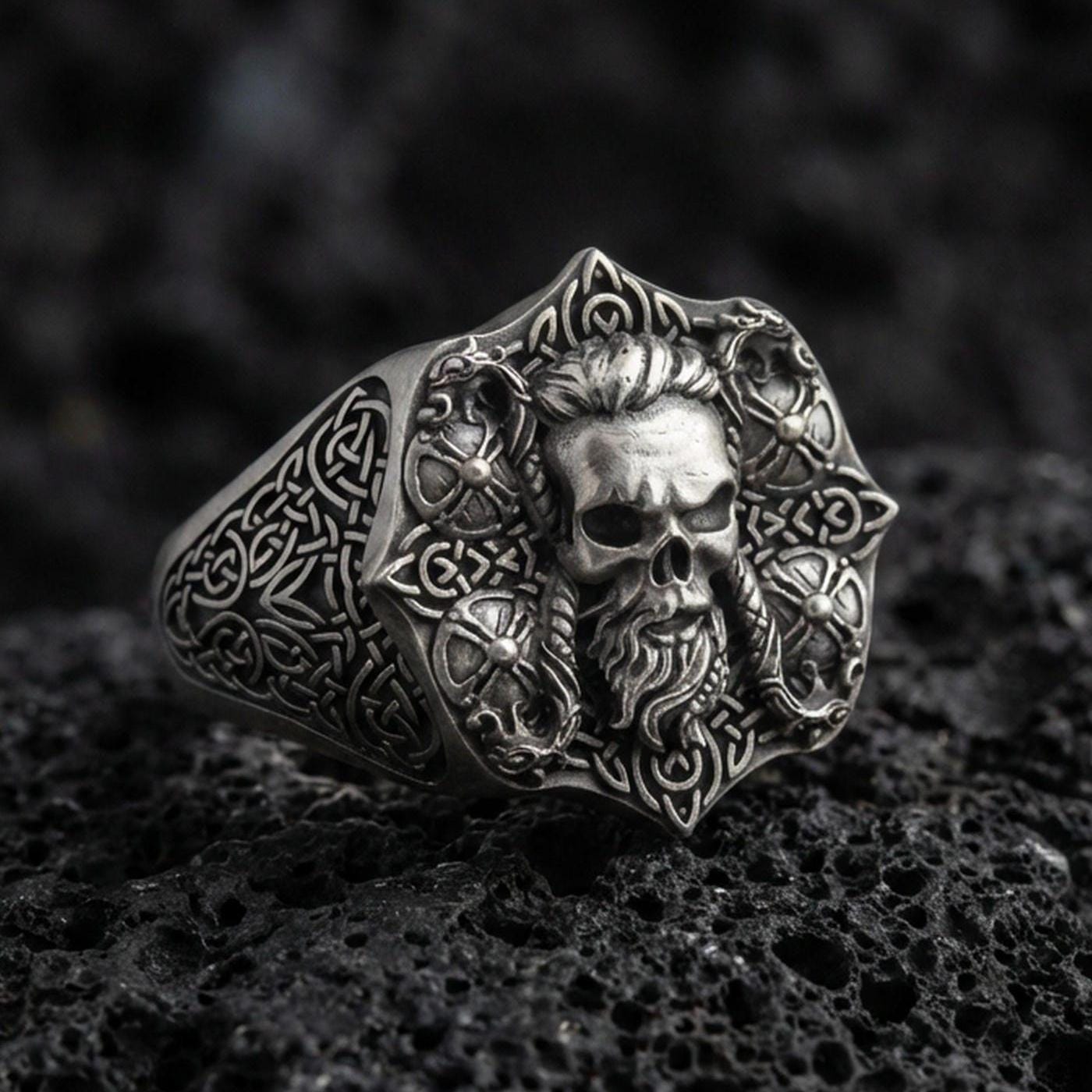 Oxidized sterling silver skull signet ring with Celtic knot engraving on black volcanic rock, macro close-up showing deep relief and antique patina