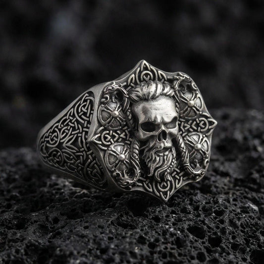 Oxidized sterling silver skull signet ring with Celtic knot engraving on black volcanic rock, macro close-up showing deep relief and antique patina