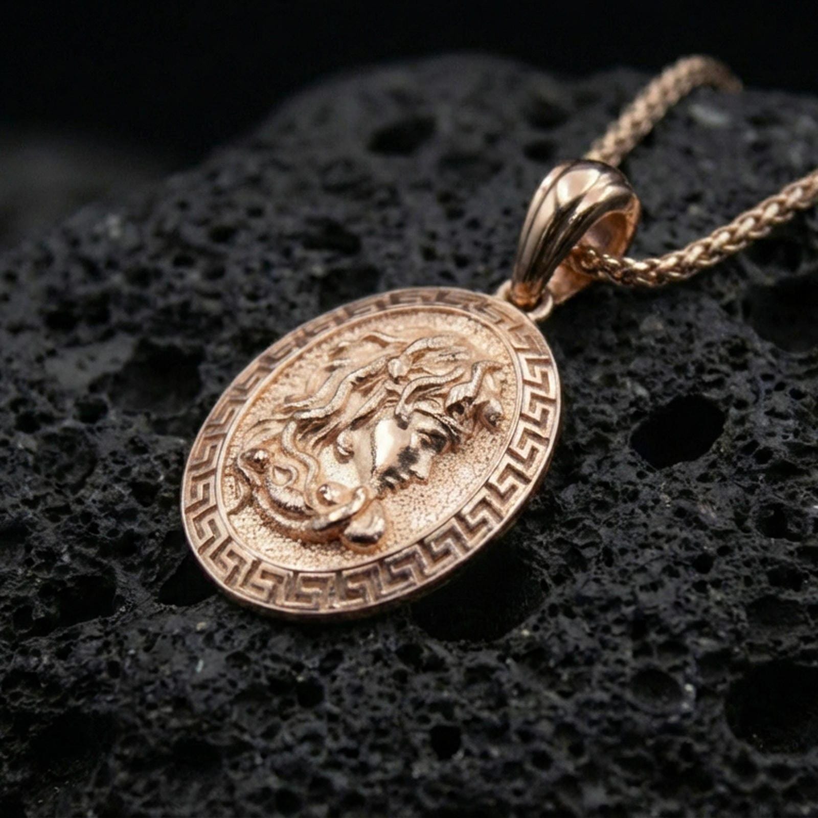Rose gold plated Medusa necklace on dark volcanic stone, Greek key coin pendant with sculpted Medusa profile and antique-inspired finish