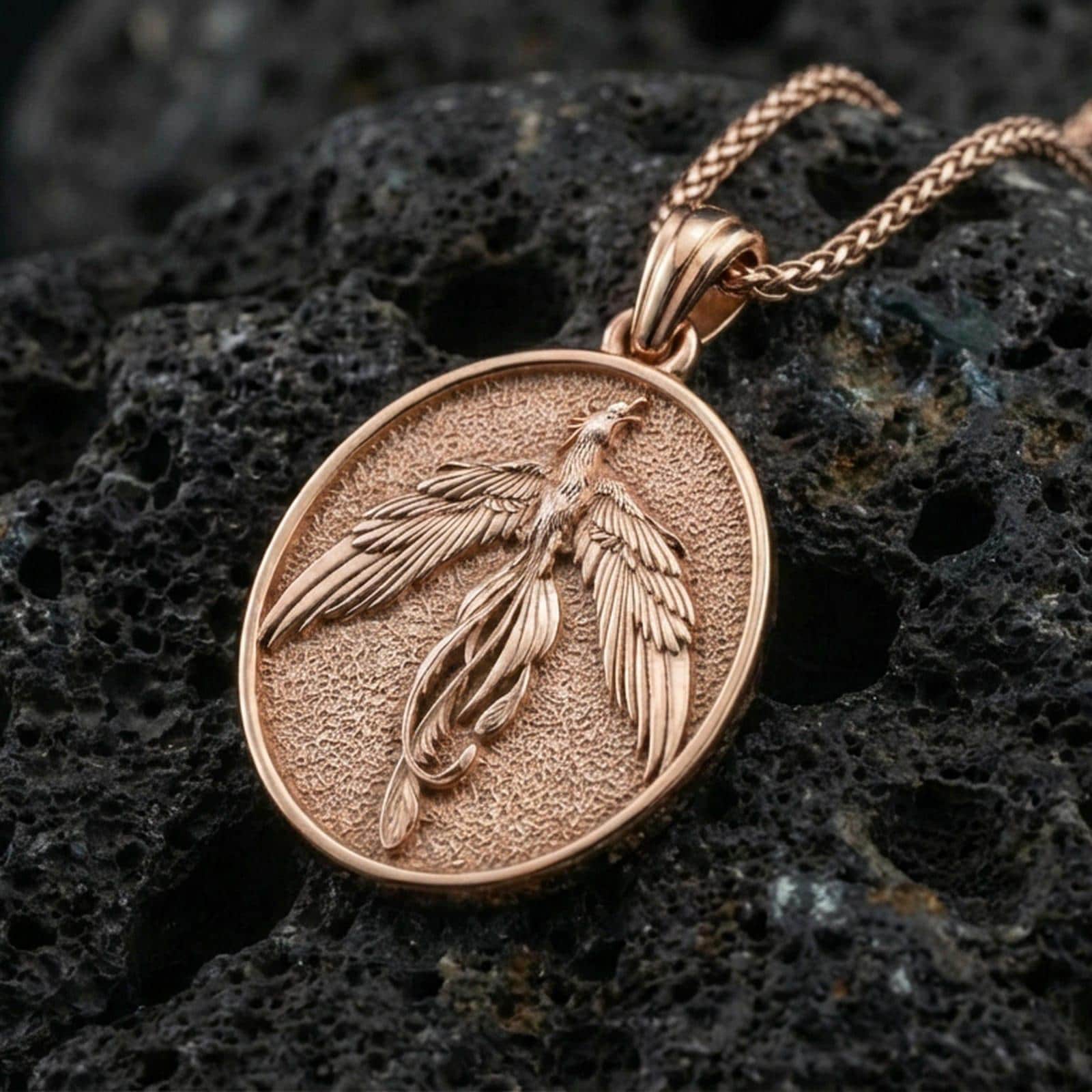 Rose gold plated phoenix medallion necklace on dark volcanic stone, elegant firebird pendant with detailed wing feathers