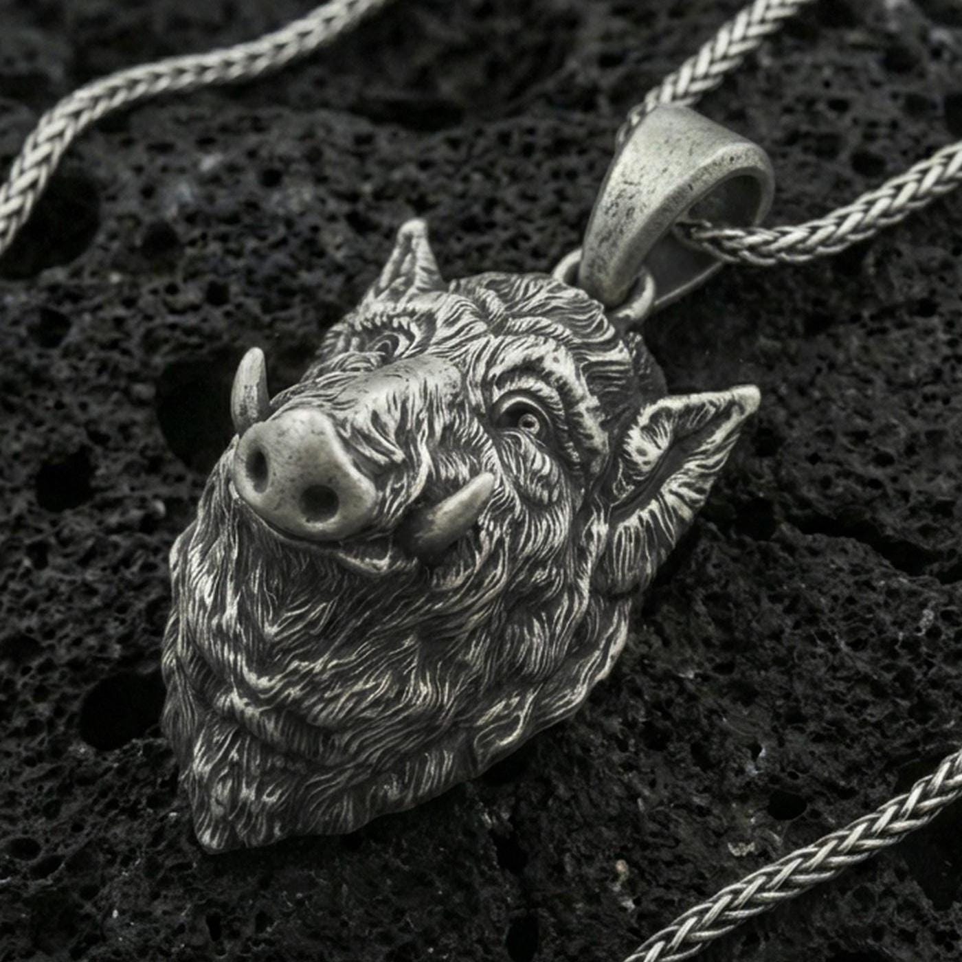 Oxidized sterling silver boar head pendant on black volcanic rock with braided chain, macro close-up showing deep fur texture and antique patina