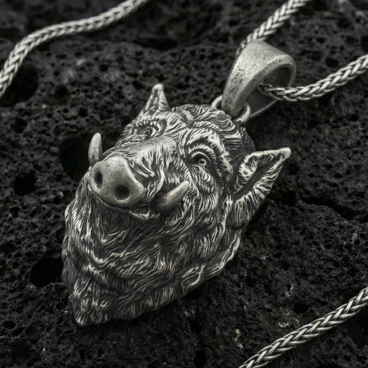 Oxidized sterling silver boar head pendant on black volcanic rock with braided chain, macro close-up showing deep fur texture and antique patina