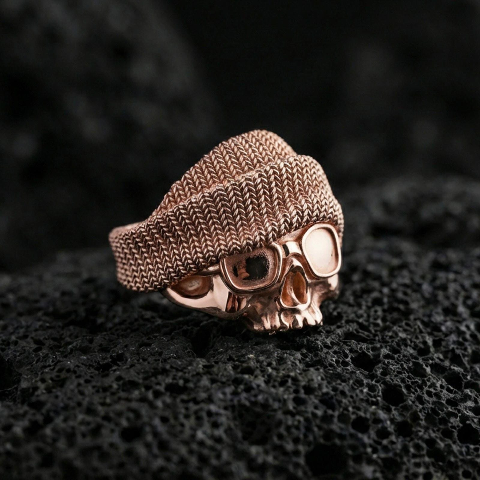 Rose gold plated skull ring with beanie and goggles on textured volcanic rock, luxury close-up capturing warm tone and sculpted beanie pattern