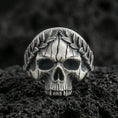 Load image into Gallery viewer, Oxidized sterling silver laurel wreath skull ring on black volcanic rock, macro close-up showing hollow eye sockets, cracked texture lines and deep relief details
