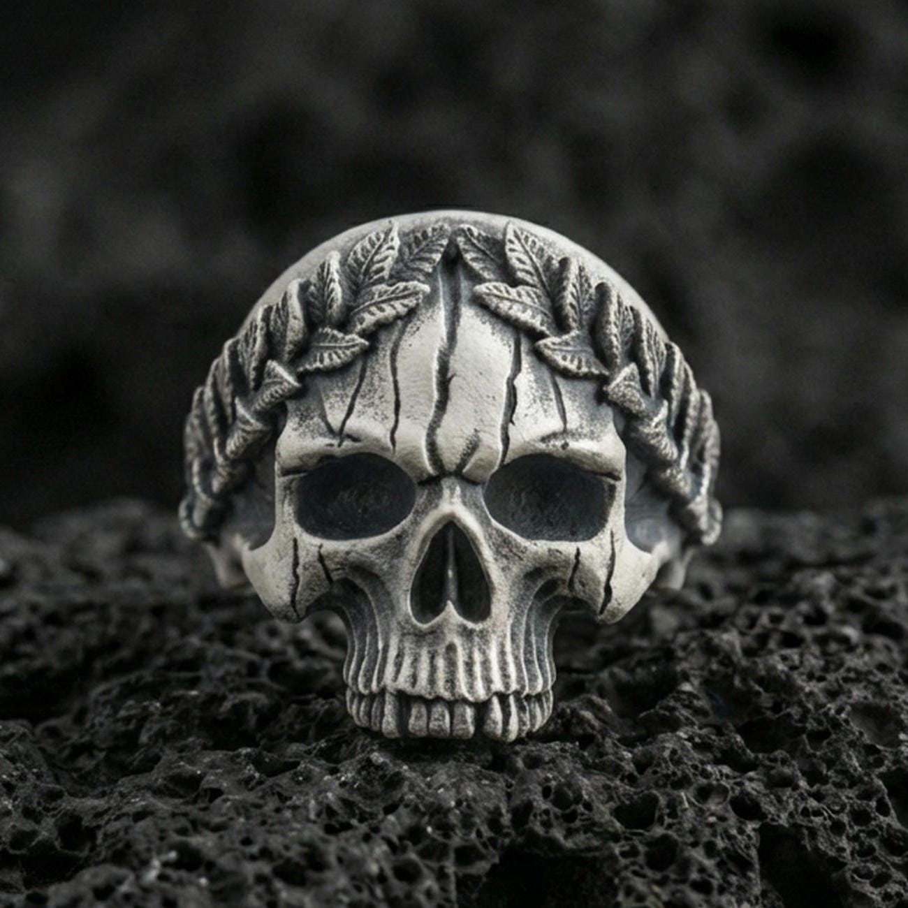 Oxidized sterling silver laurel wreath skull ring on black volcanic rock, macro close-up showing hollow eye sockets, cracked texture lines and deep relief details