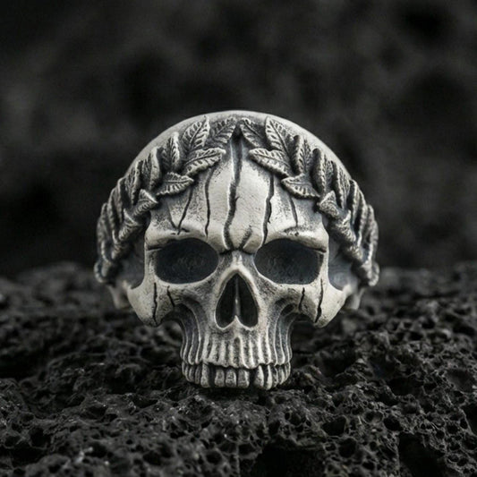 Oxidized sterling silver laurel wreath skull ring on black volcanic rock, macro close-up showing hollow eye sockets, cracked texture lines and deep relief details