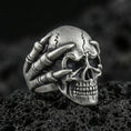 Load image into Gallery viewer, Oxidized sterling silver skull biker ring with skeletal claw hand gripping the skull on black volcanic rock, macro close-up showing deep shadows and gothic texture
