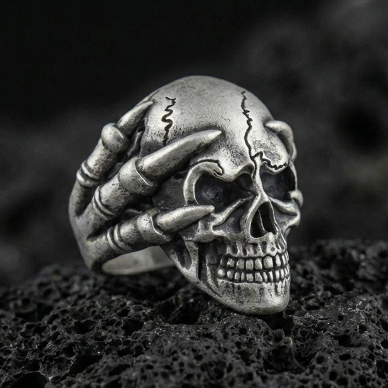 Oxidized sterling silver skull biker ring with skeletal claw hand gripping the skull on black volcanic rock, macro close-up showing deep shadows and gothic texture