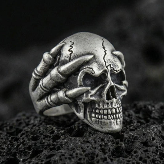 Oxidized sterling silver skull biker ring with skeletal claw hand gripping the skull on black volcanic rock, macro close-up showing deep shadows and gothic texture
