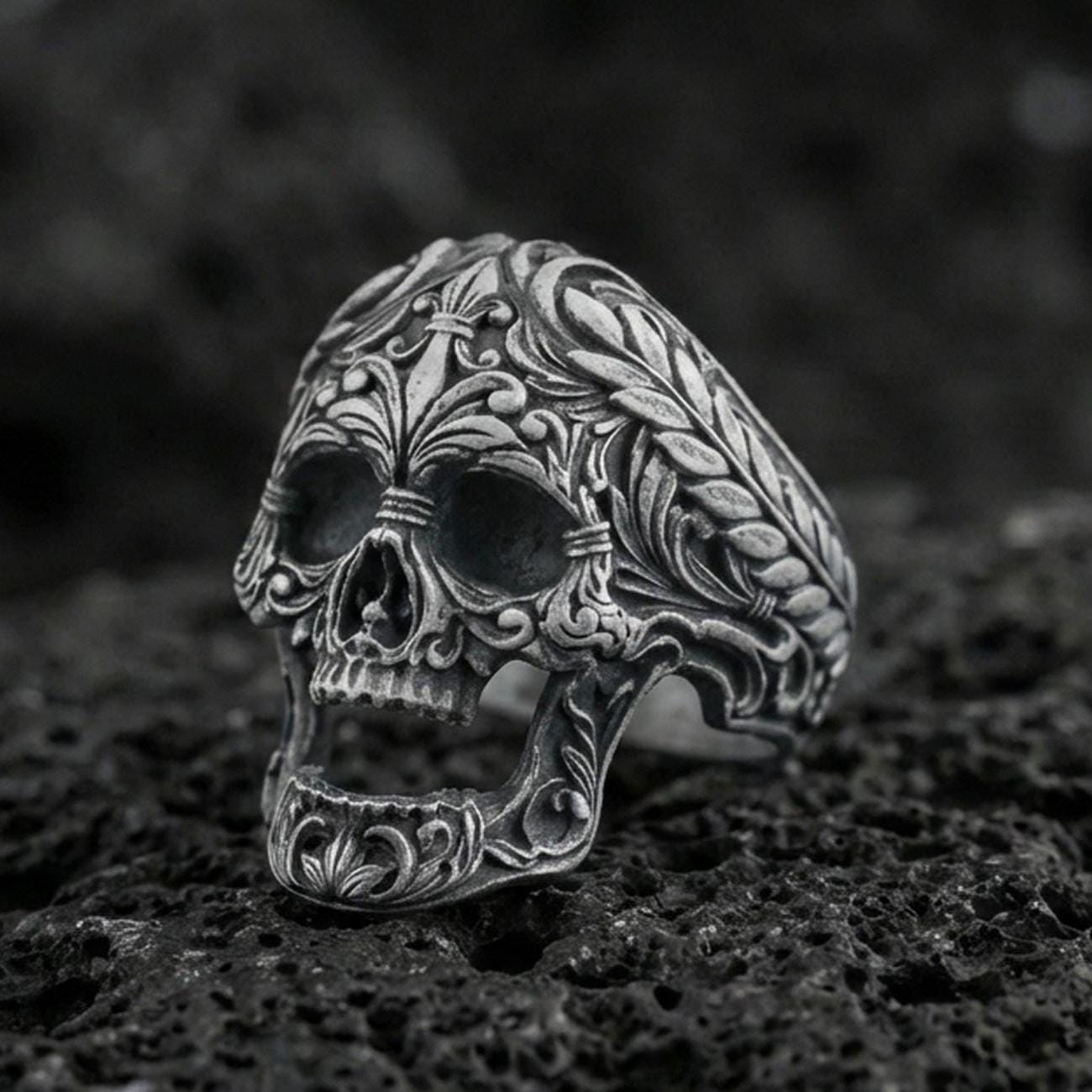 Oxidized sterling silver baroque skull ring with ornate floral filigree and braided border on black volcanic rock, macro close-up with deep shadowed detail