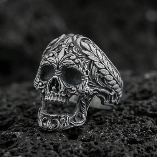 Oxidized sterling silver baroque skull ring with ornate floral filigree and braided border on black volcanic rock, macro close-up with deep shadowed detail