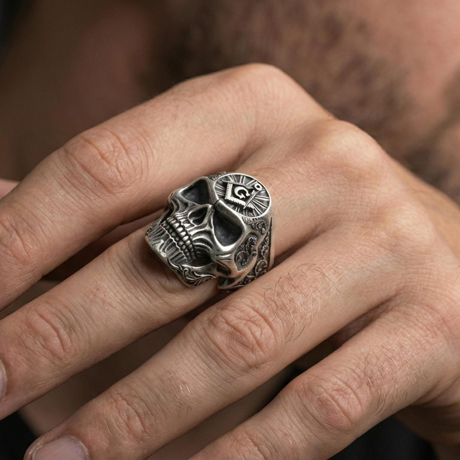 Oxidized skull ring with square and compass “G” emblem worn on a finger, close-up lifestyle photo highlighting deep shadowed relief and engraved sides