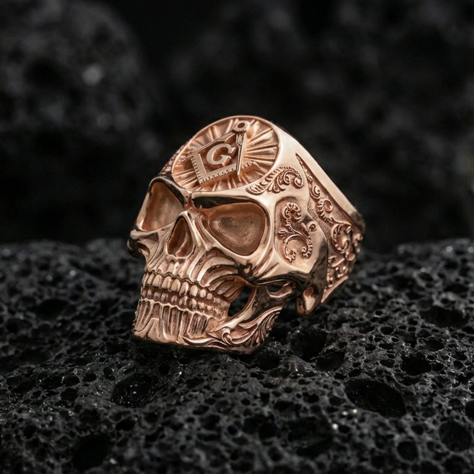 Rose gold plated skull ring with square and compass “G” symbol on the crown and detailed scrollwork band, shot on black volcanic rock