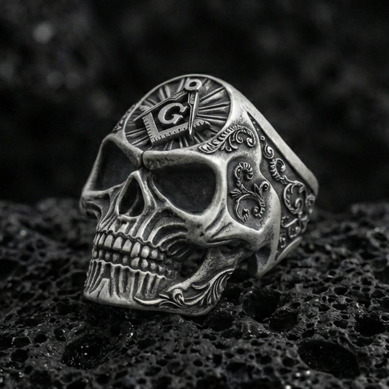 Oxidized sterling silver skull ring with Masonic square and compass “G” emblem on the crown, on black volcanic rock in a dark macro studio shot