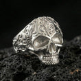 Load image into Gallery viewer, Polished silver skull ring with detailed floral scrollwork band and bright highlights, macro close-up on textured volcanic stone

