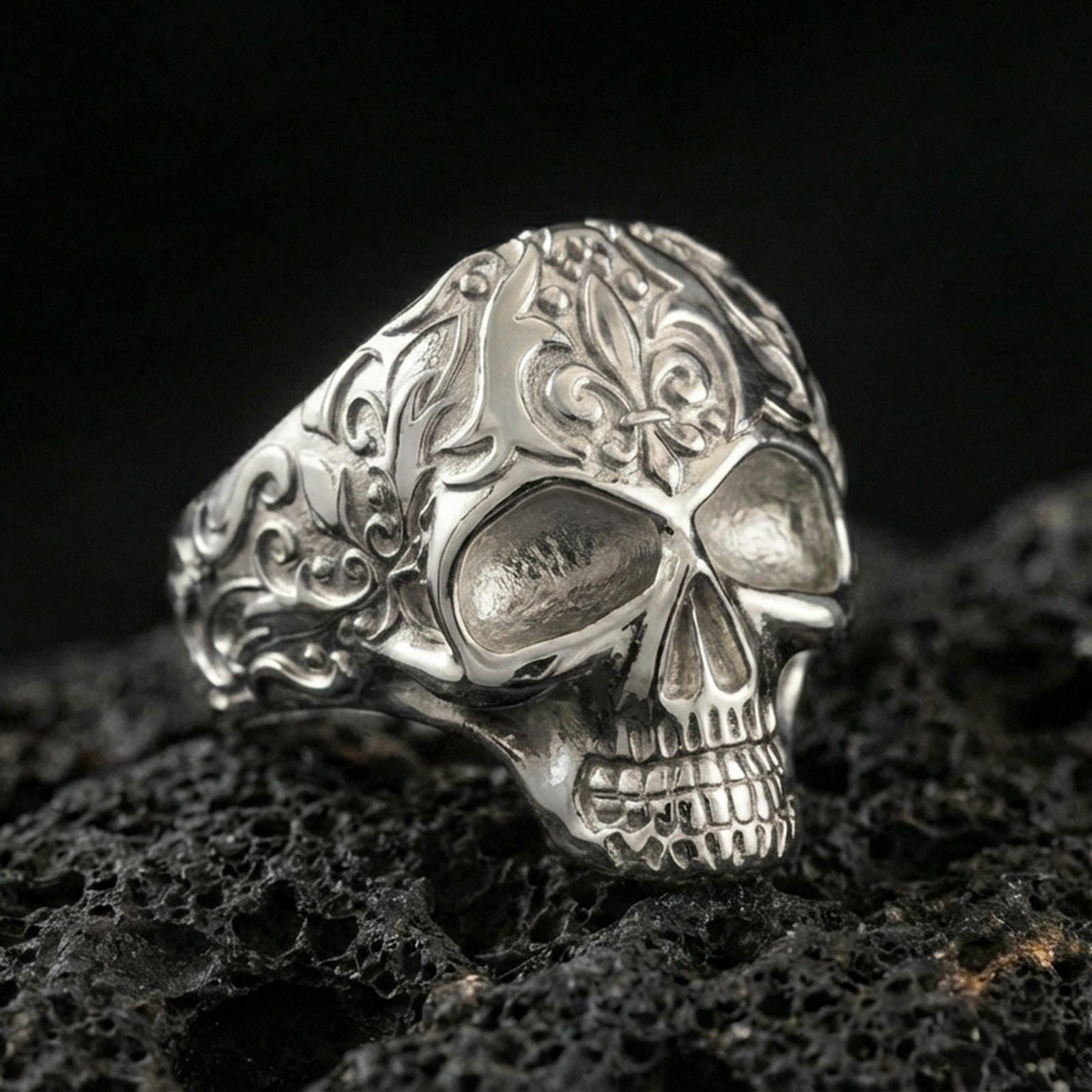 Polished silver skull ring with detailed floral scrollwork band and bright highlights, macro close-up on textured volcanic stone