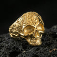 Load image into Gallery viewer, Gold plated ornate skull ring with baroque-style filigree engraving on the band, product photo on porous volcanic rock under dramatic lighting
